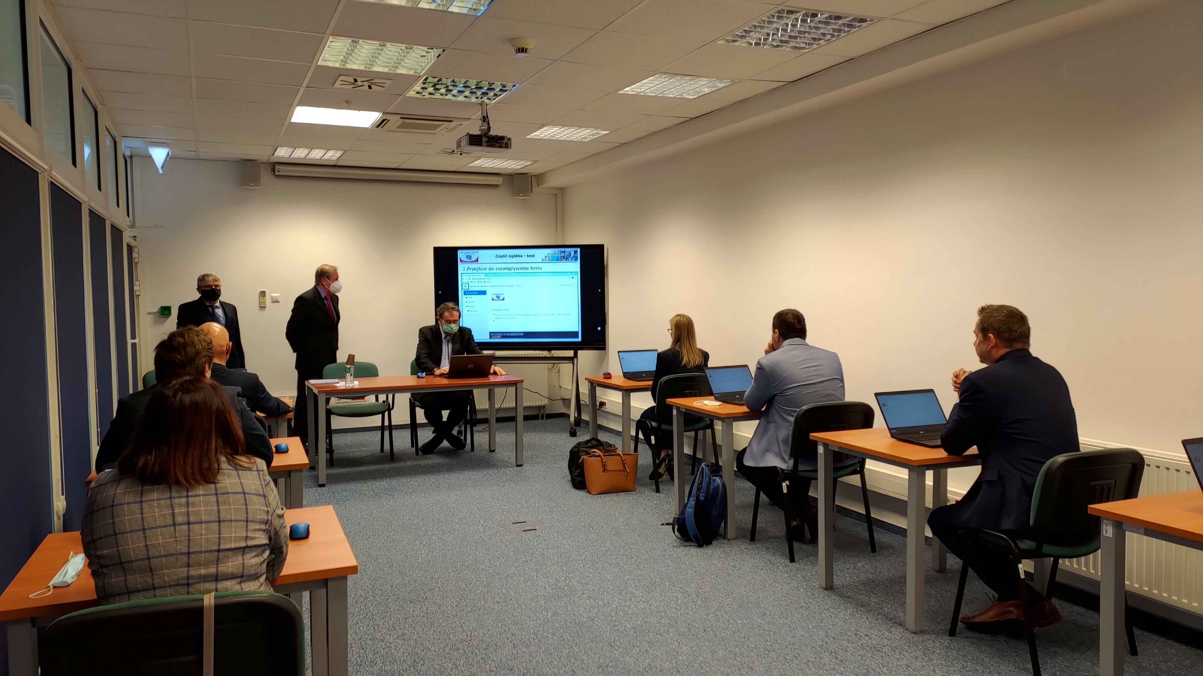 Zdjęcie przedstawia salę egzaminacyjną z członkami Komisji egzaminacyjnej i osobami przystępującymi do egzaminu siedzącymi pojedynczo przed laptopami