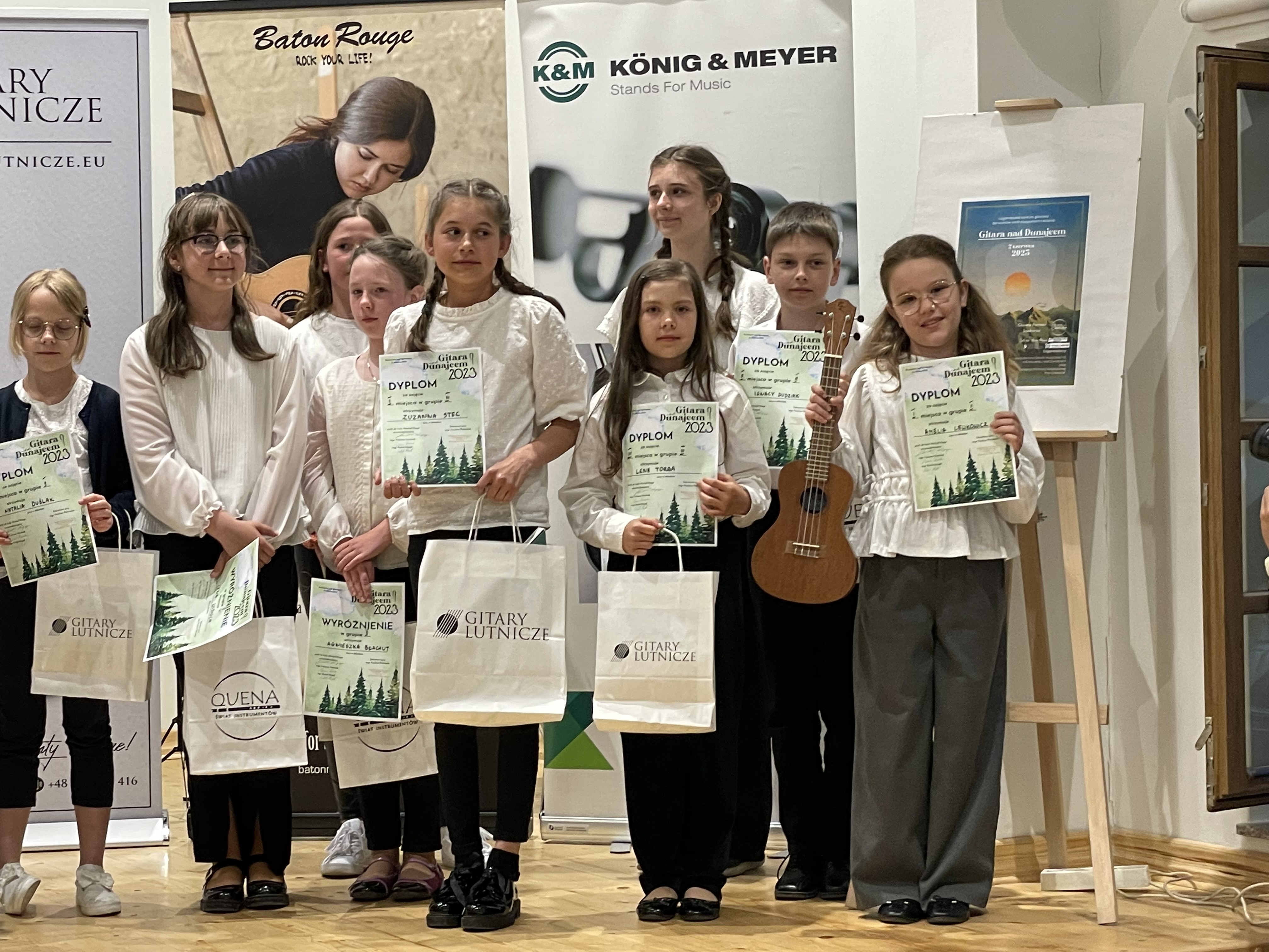  Stoi na scenie wraz z innymi laureatami konkursu, trzyma w dłoni dyplom i torbę z nagrodami. Inne dzieci również trzymają dyplomy i nagrody w torbach.