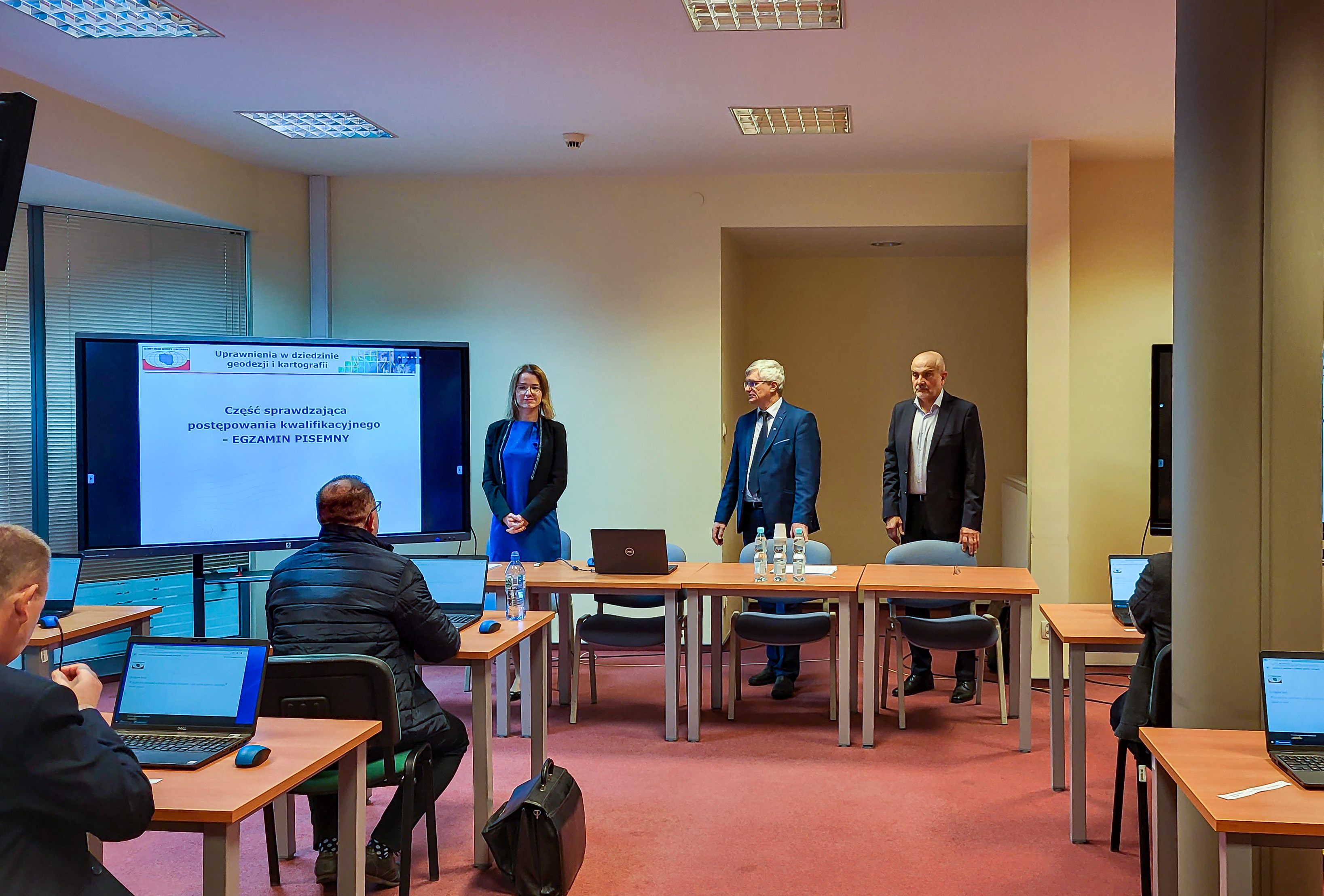 Zdjęcie przedstawia salę egzaminacyjną z członkami Komisji egzaminacyjnej i osobami przystępującymi do egzaminu siedzącymi pojedynczo przed laptopami.