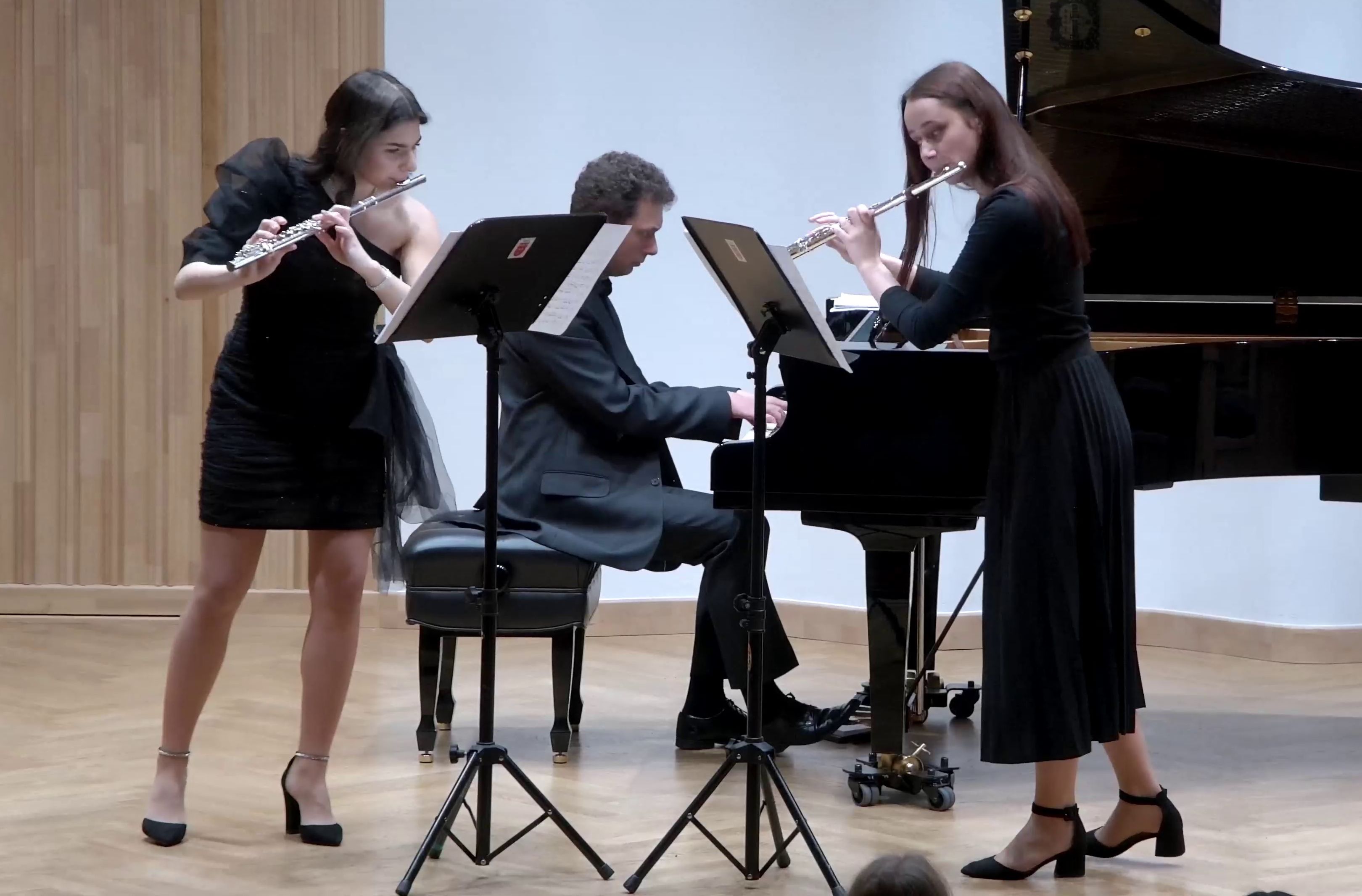 Zdjęcie. na estradzie sali koncertowej występują dwie flecistki, którym towarzyszy pianista, grający na dużym czarnym fortepianie. Przed każdą flecistką stoi pulpit z nutami.