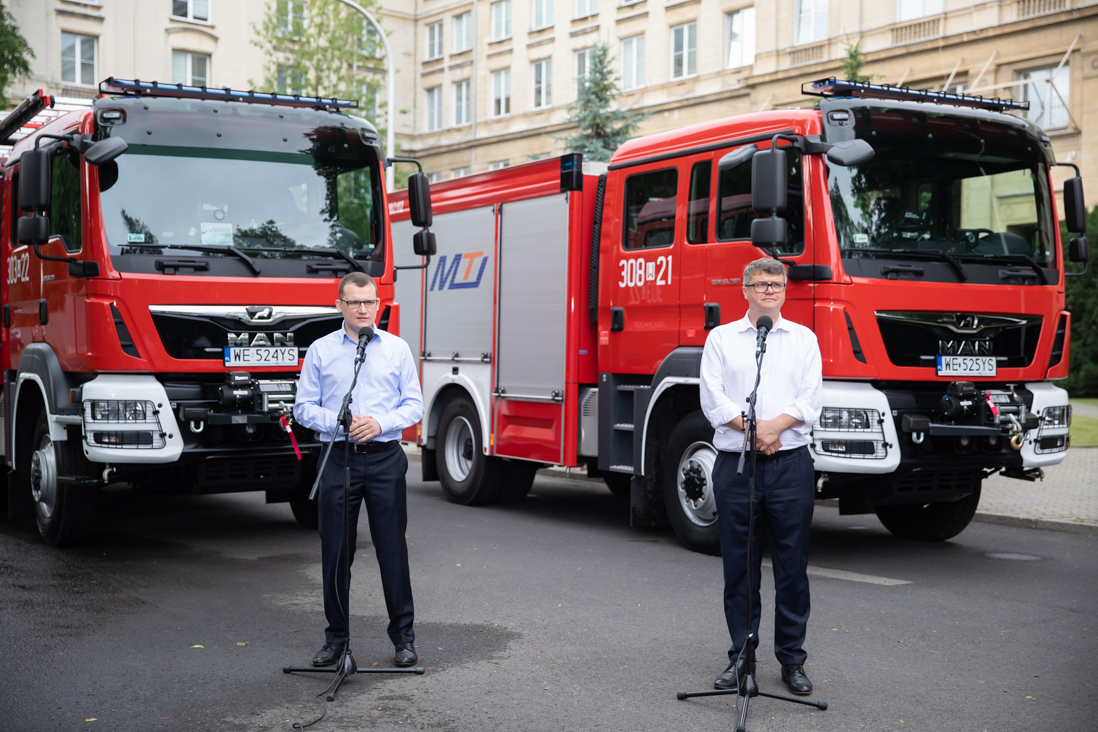 Na zdjęciu widać stojących przy mikrofonach Pawła Szefernakera wiceministra SWiA (przemawia) i Macieja Wąsika wiceministra MSWiA. W tle widać dwa stojące, ciężarowe samochody Straży Pożarnej.