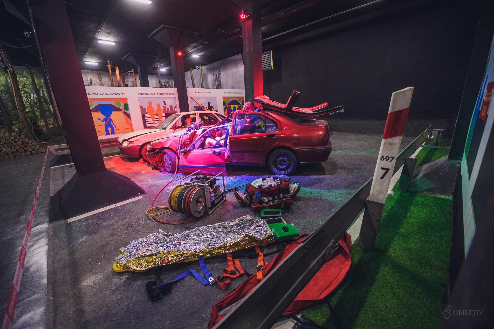 Zdjęcie przedstawia pozorację wypadku samochodowego. Dwa rozbite samochody oraz sprzęt do ratownictwa drogowego i medycznego.