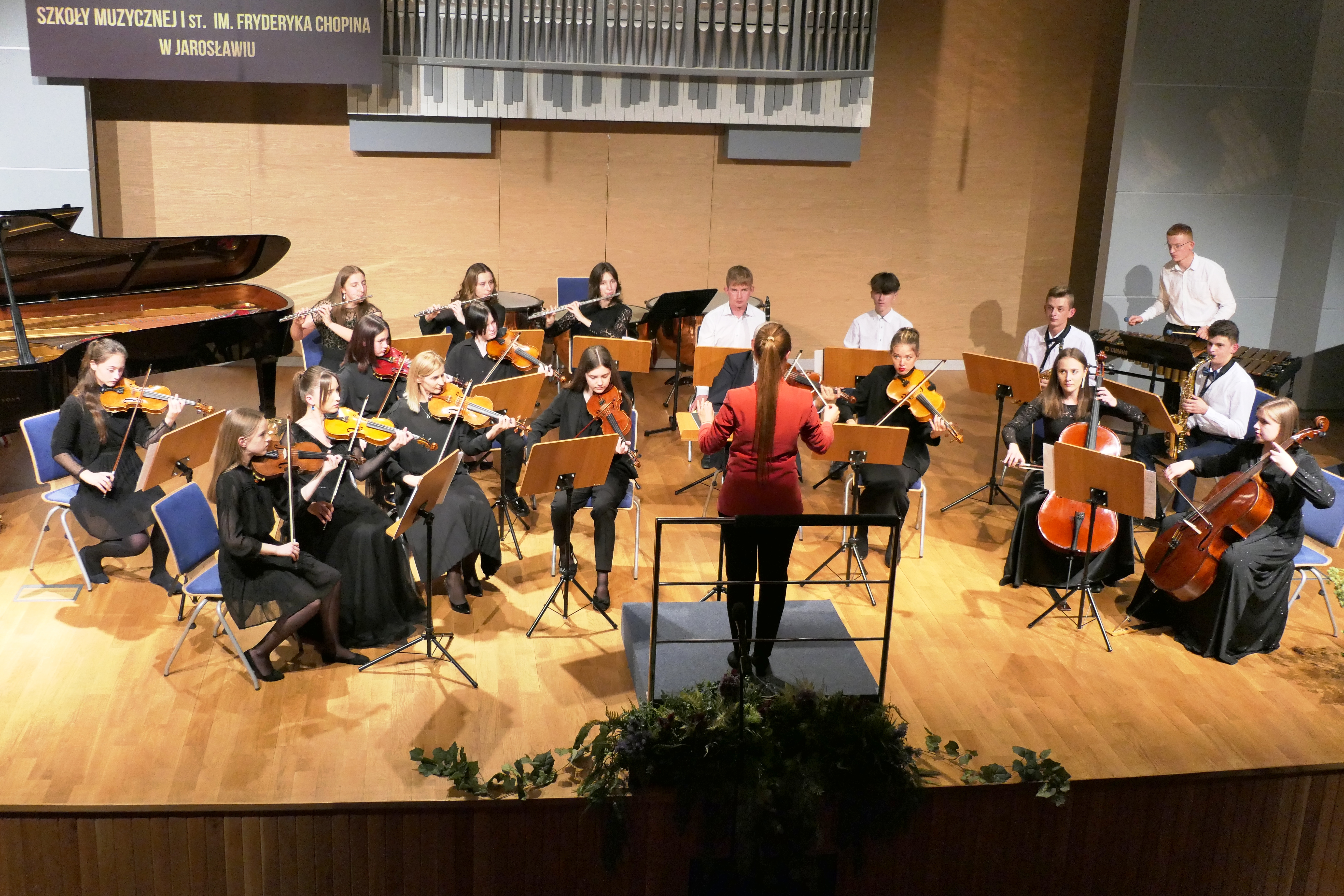 Zdjęcie występu orkiestry Szkoły Muzycznej II st. w Jarosławiu. Na scenie od lewej strony uczniowie grający na skrzypcach za nimi na fletach, na środku na altówkach za nimi osoby grające na klarnecie i trąbce, po lewej na wiolonczeli, za nimi na saksofonach, a za saksofonami uczeń grający na wibrafonie