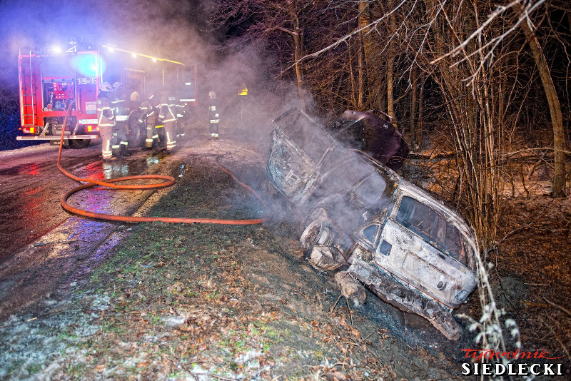 Pożar samochodu osobowego, który znajduje się w rowie obok jezdni. Na zdjęciu spalony wrak pojazdu, w tle strażacy.