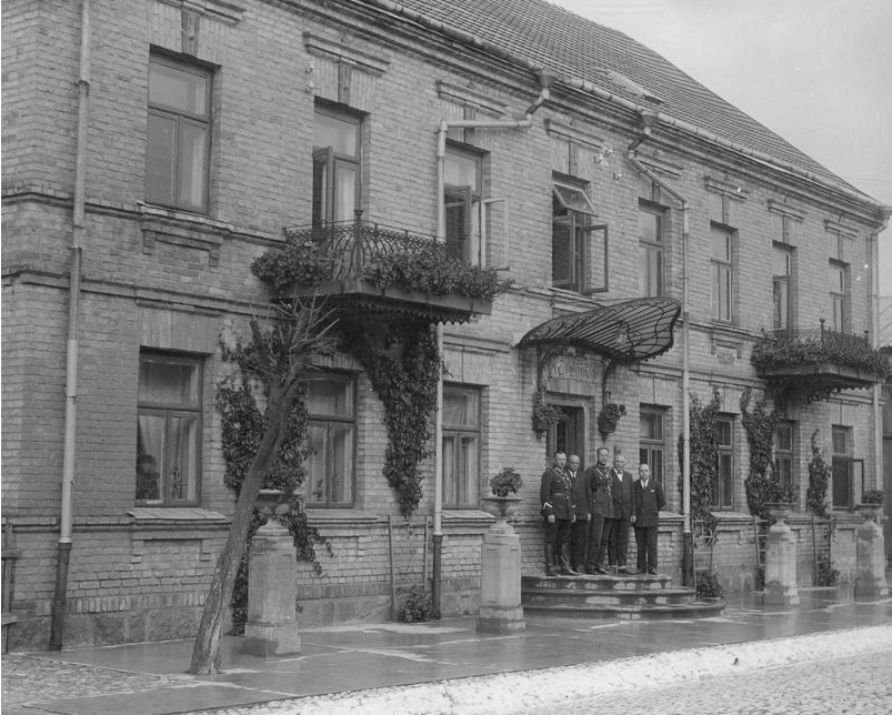 Czarno-biały widok dawnego budynku szkoły. Przed wejściem pięciu mężczyzn w garniturach i mundurach.