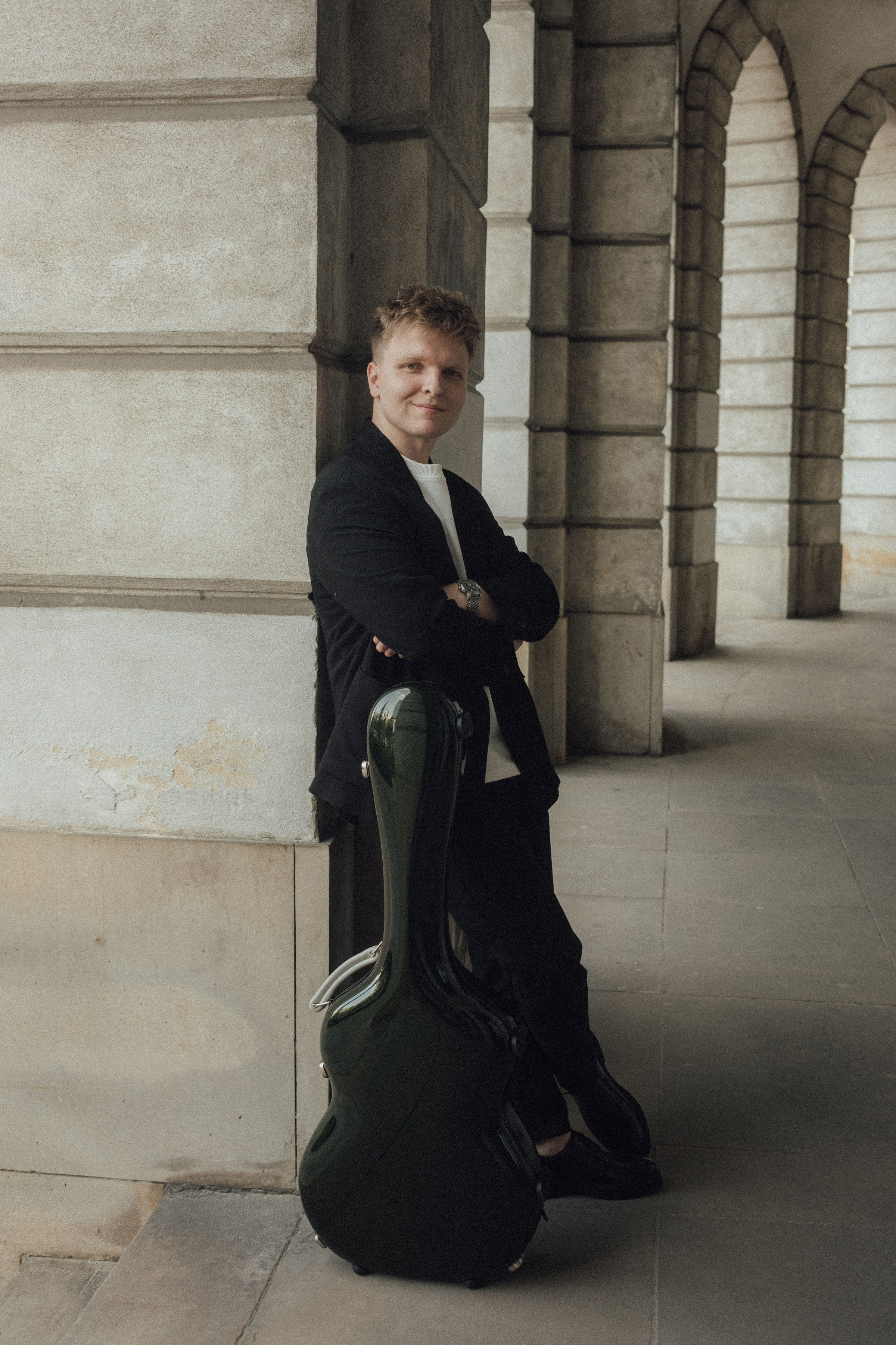Zdjęcie przedstawiające artystę opartego o kolumnę z futerałem na gitarę na tle arkad.