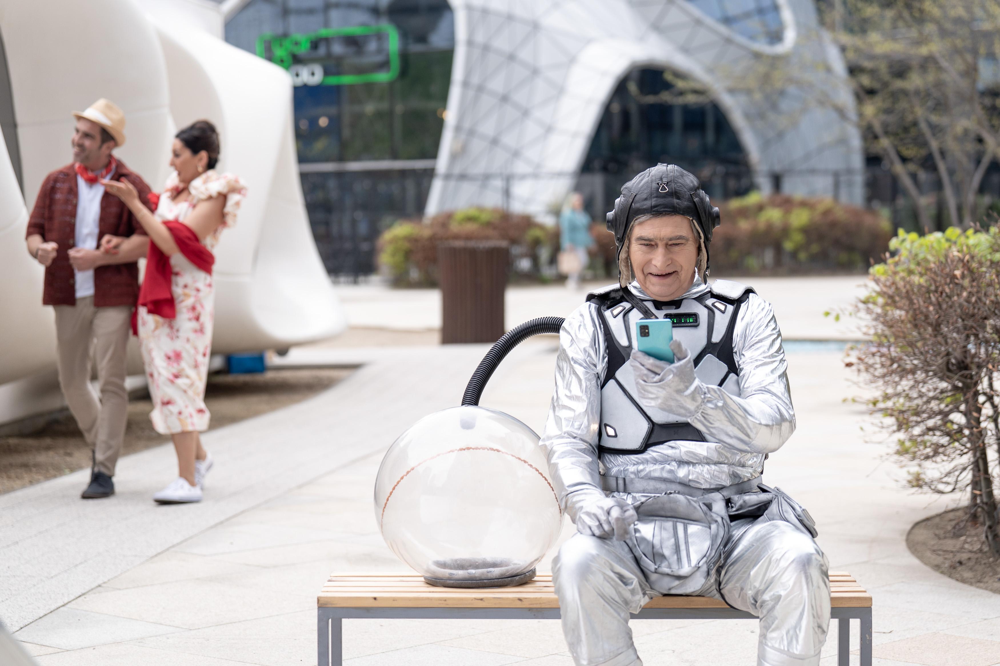 Uśmiechnięty mężczyzna w stroju astronauty siedzi na ławce i patrzy w telefon komórkowy, obok niego leży hełm; w tle po lewej stronie stoją mężczyzna i kobieta, którzy coś oglądają