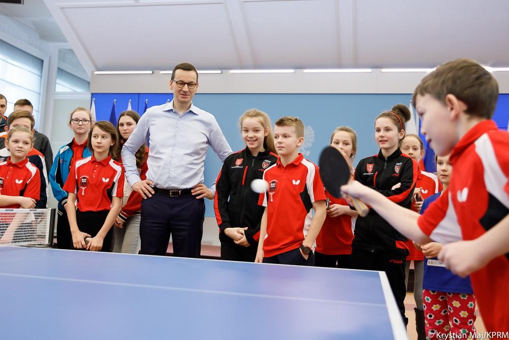 Premier Mateusz Morawiecki stoi wśród dzieci, a przed nimi chłopiec gra w ping-ponga.
