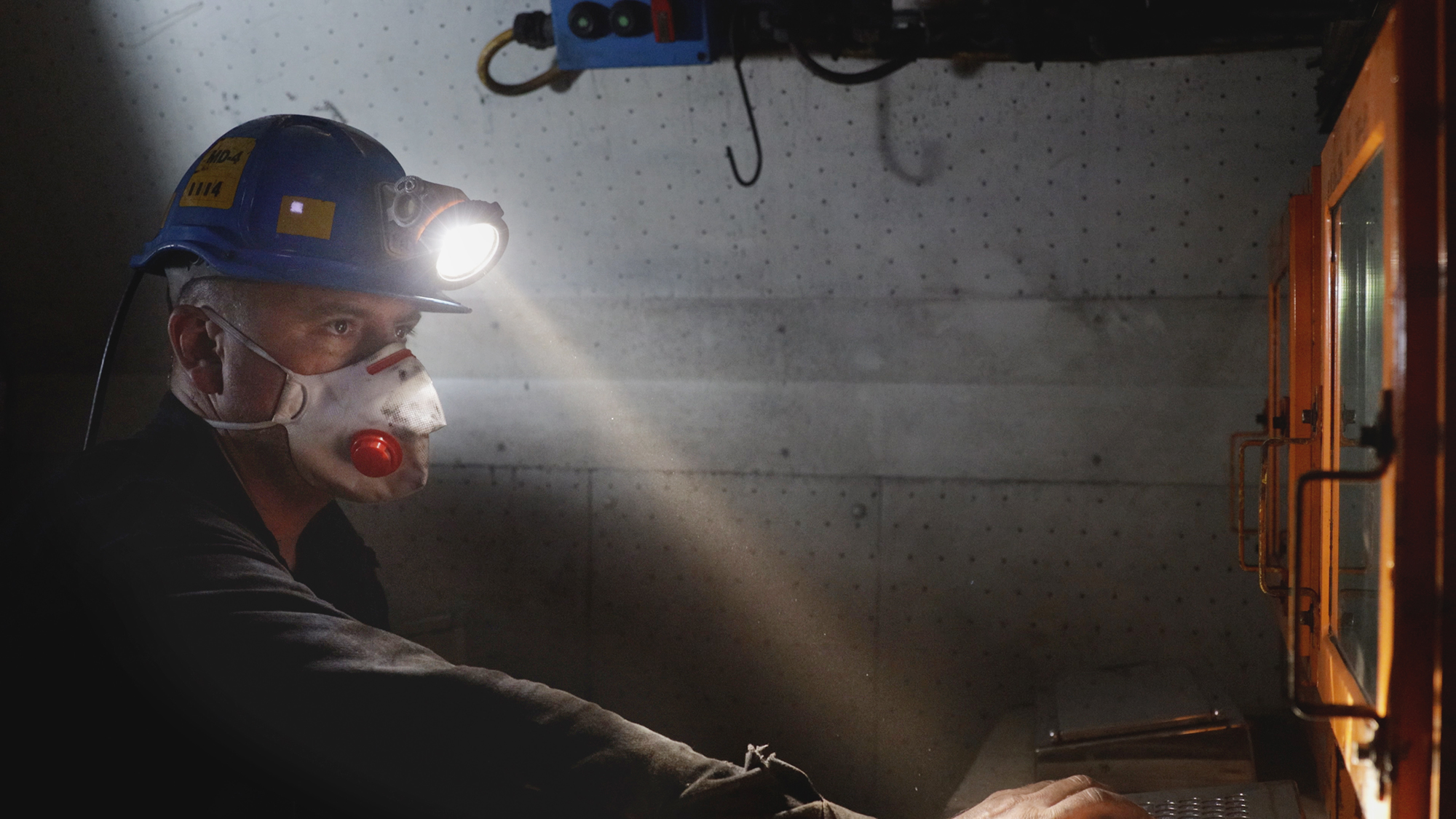 Mężczyzna w czarnym stroju i niebieskim kasku, w maseczce obsługuje specjalistyczny sprzęt. Rękę trzyma na klawiaturze i patrzy na wyświetlacz