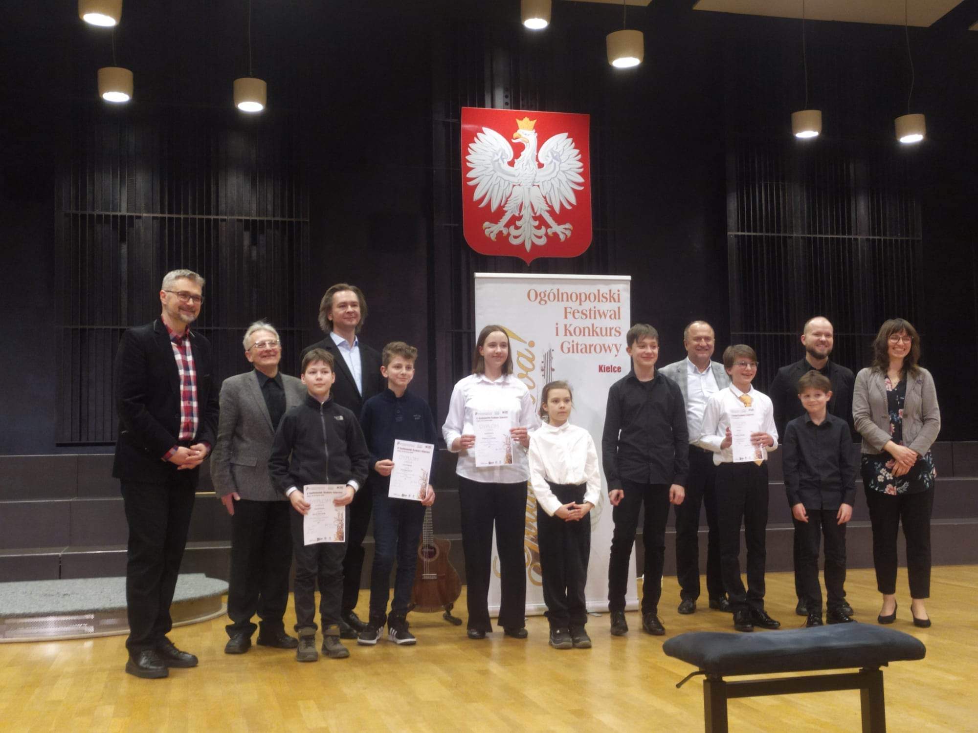 Sala koncertowa Zespołu Szkół Muzycznych w Kielcach, ogromna scena. Na środku sceny stoją laureaci X Ogólnopolskiego Festiwalu i Konkursu "Gitara Viva" w dniu 16 marca 2023 r. Między nimi, po środku stoi uczennica naszej szkoły Lena Torba - najmniejsza ze wszystkich osób, która otrzymała wyróżnienie. Po środku za nimi na ścianie wisi duże godło Polski, pod nim stoi baner z napisem "Gitara Viva!", na suficie wisi mnóstwo żyrandoli. Wśród laureatów stoją jurorzy konkursu, w sumie stoi 13 osób.