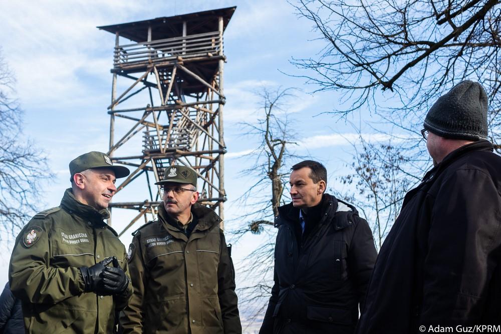 Premier Mateusz Morawiecki i strażnicy graniczni stoją pod wieżą obserwacyjną.