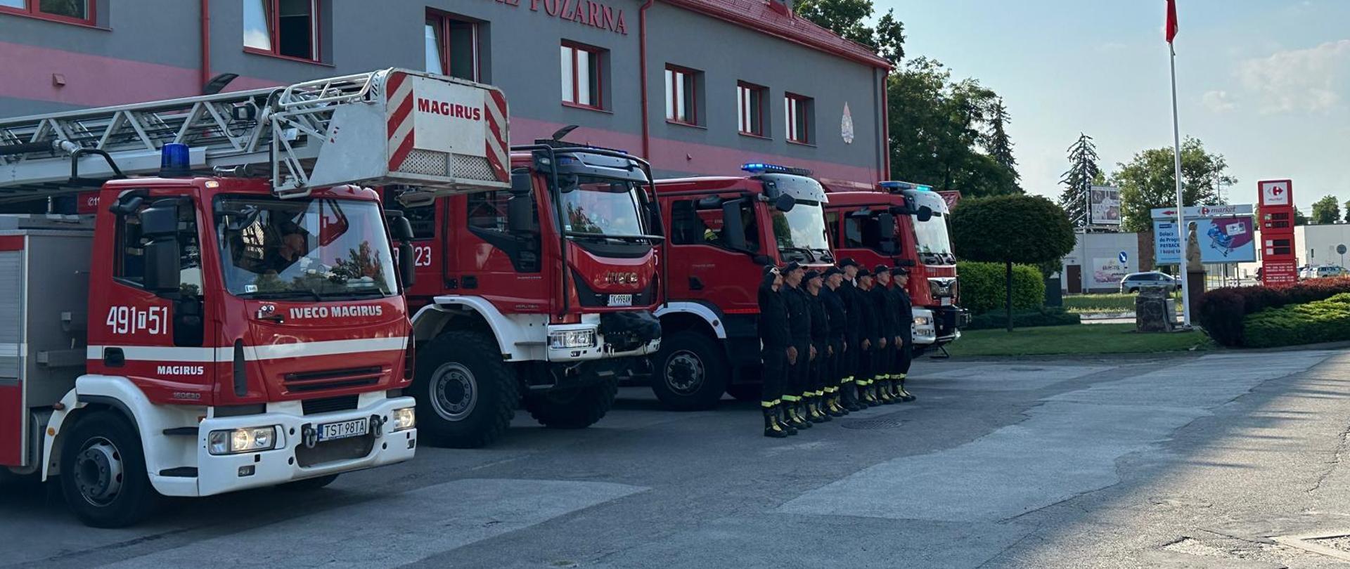 Strażacy w szyku oddający honory w tle pojazdy pożarnicze