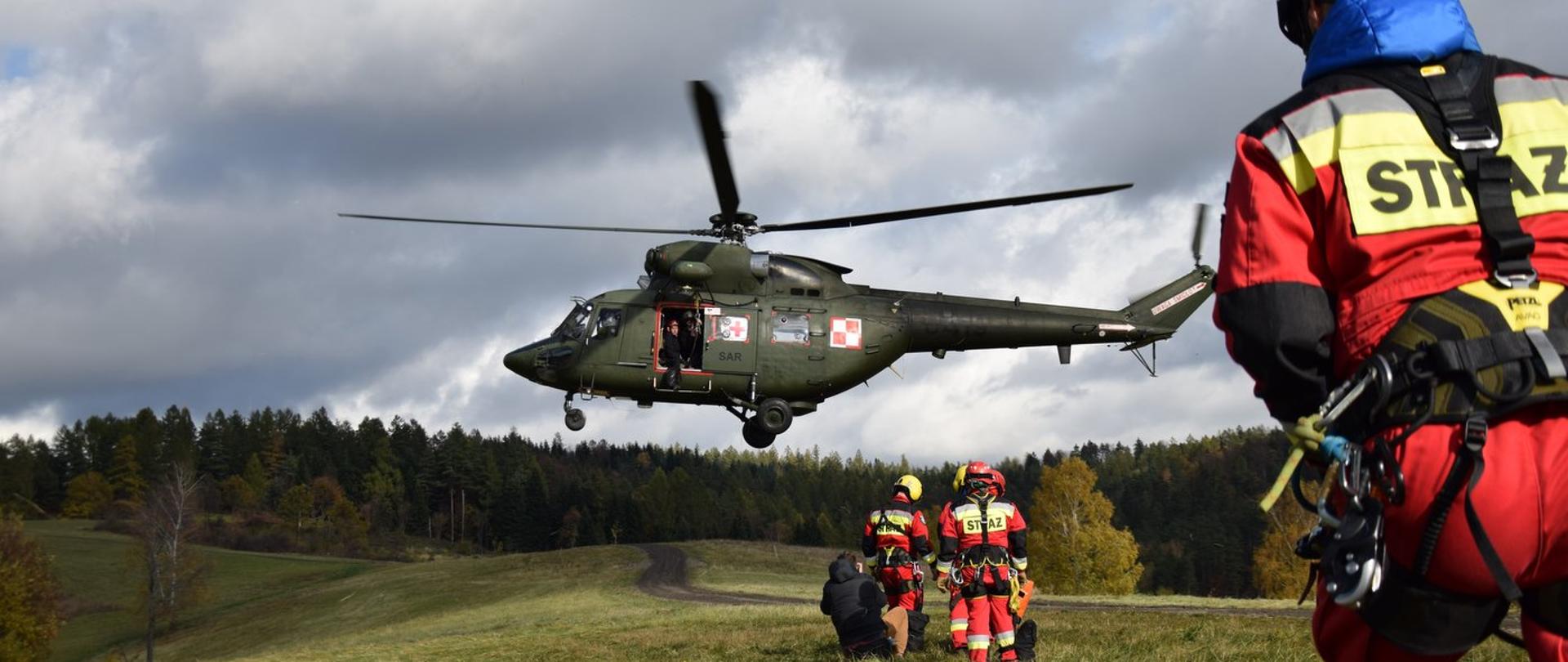 Zdjęcie zrobione na zewnątrz, podczas manewrów. W powietrzu śmigłowiec SAR, Na ziemi stoją strażacy, którzy przygotowują się do wejścia na pokład śmigłowca