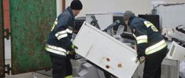Strażacy dla Tosi. Plac komendy w Rawiczu. Na nim złożone są elektrośmieci w postaci m.in lodówek, monitorów komputerowych, drukarek, itp. Dwóch strażaków przenosi lodówkę do podstawionego kontenera.