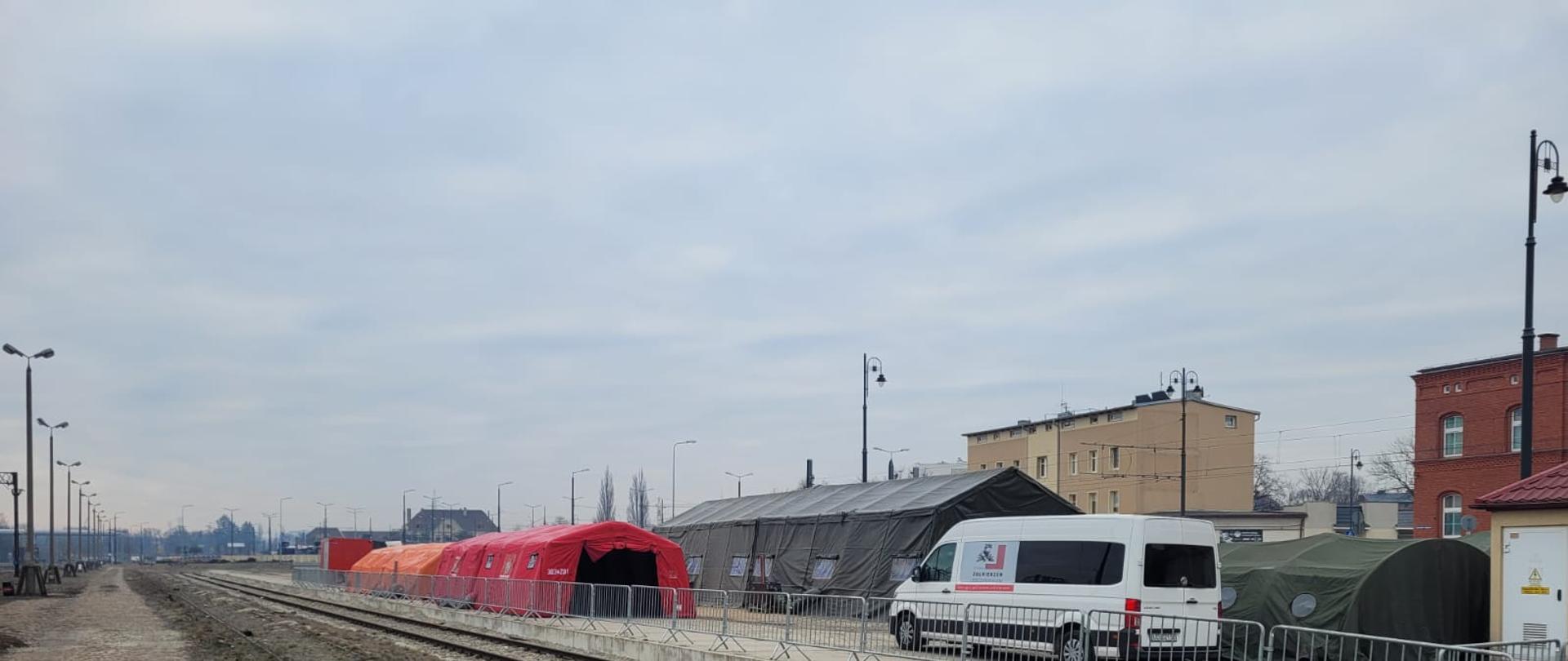 Perony dworca, na których stoją z lewej czerwone i pomarańczowe namioty strażackie, z lewej zielone namioty wojskowe. W środku widać białego busa. 