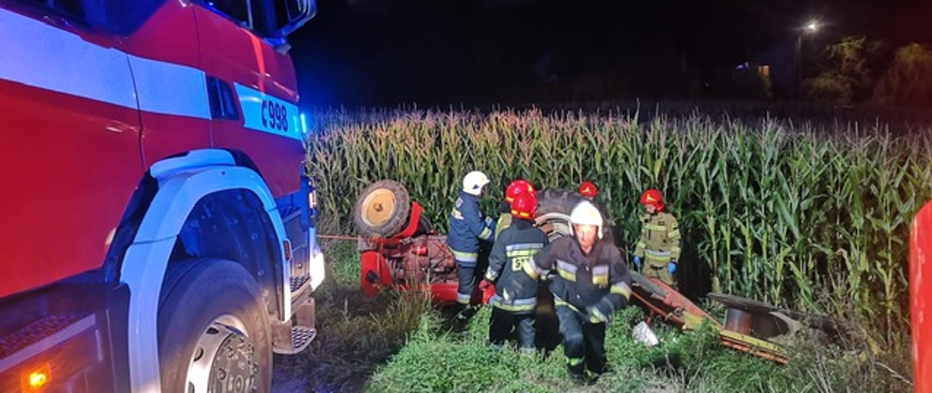 Na zdjęciu widoczny samochód pożarniczy oraz strażacy przy przewróconym ciągniku rolniczym.