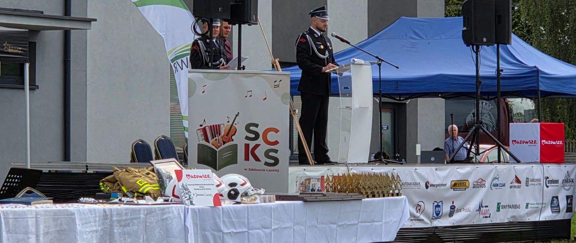 Na zdjęciu widoczna jest oficjalna trybuna ustawiona przy nowoczesnym budynku urzędu/ośrodka kultury. Druh w mundurze galowym wygłasza przemówienie przy mównicy z nagłośnieniem.
Stół z odznaczeniami i pucharami: Na pierwszym planie zdjęcia znajduje się nakryty białym obrusem stół, na którym przygotowano medale, odznaczenia, okolicznościowe statuetki, dyplomy oraz strażackie hełmy paradne przeznaczone do wręczenia wyróżnionym strażakom i gościom. Banery oraz mównica nawiązują do wsparcia wydarzenia przez Samorząd Województwa Mazowieckiego.