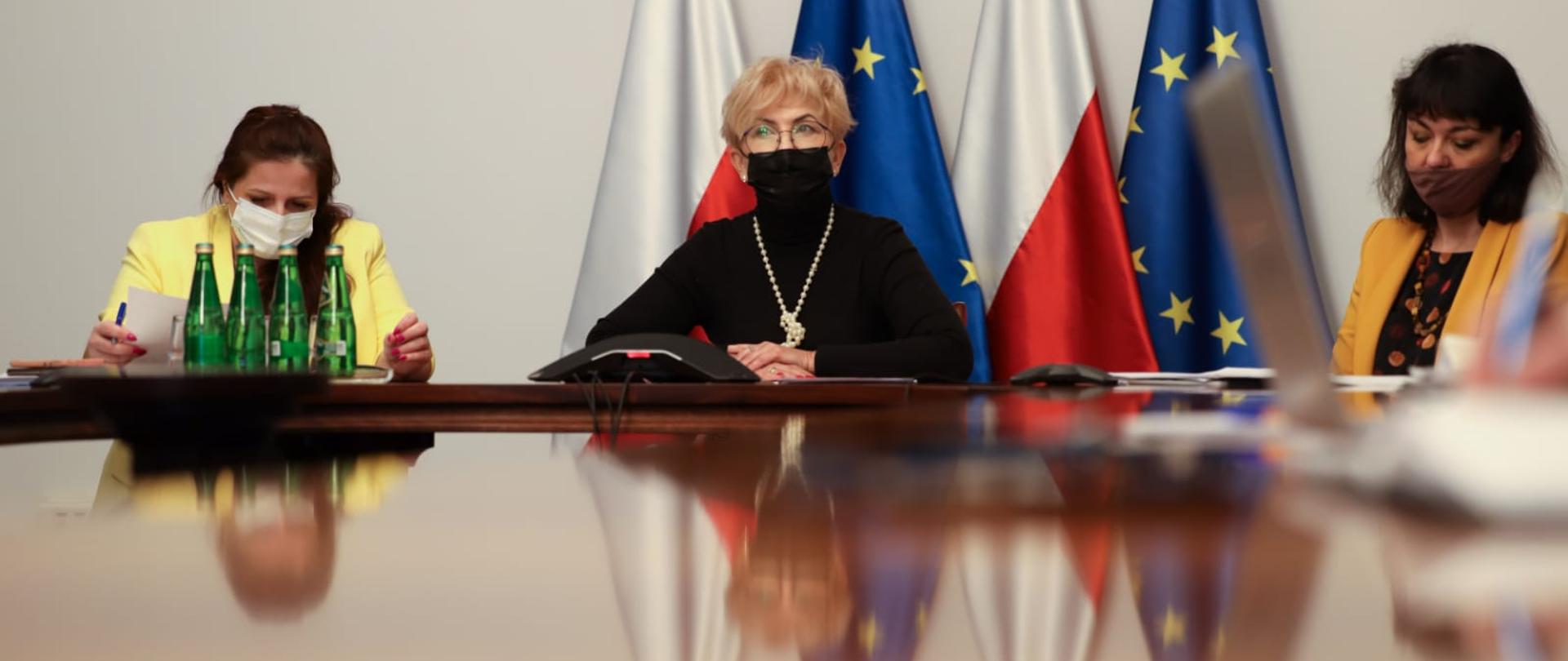 Iwona Michałek siedzi przy stole konferencyjnym. Na twarzy ma maseczkę. Przed sobą trzyma notatki. Obok siedzą osoby z departamentów merytorycznych.