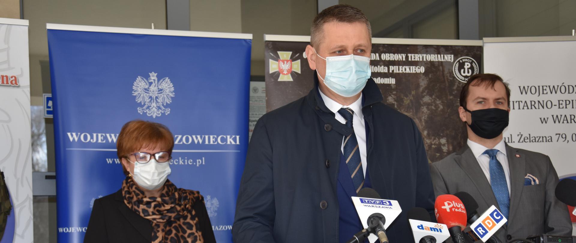 Zdjęcie z briefingu prasowego. Na pierwszym planie Wicewojewoda stojący przy mównicy, na której postawione są mikrofony dziennikarzy. W tle rollupy, przy których stoją inni uczestnicy briefingu.
