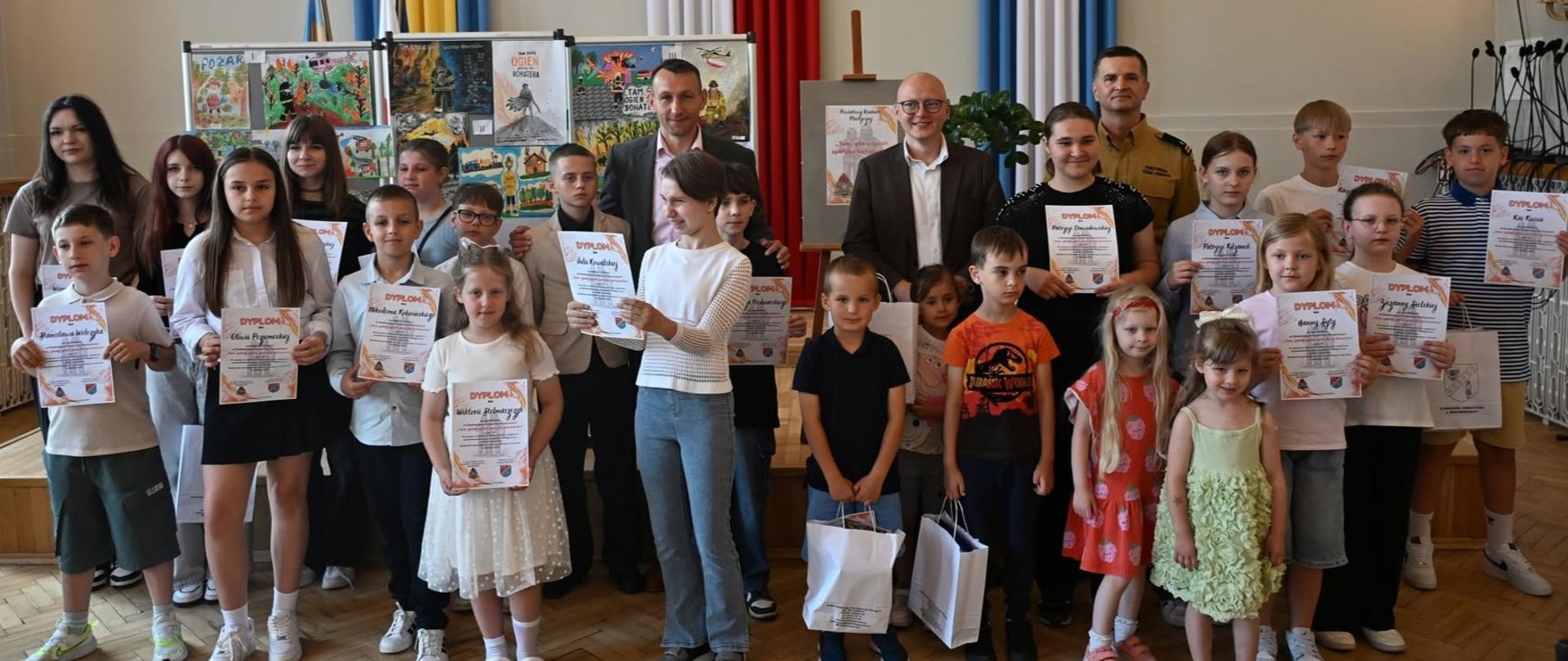 Zdjęcie grupowe w sali oficjalnej. Duża grupa dzieci i młodzieży w różnym wieku stoi w dwóch rzędach i pozuje do zdjęcia, trzymając w rękach pamiątkowe dyplomy oraz białe upominki w torbach. W centralnej części, z tyłu, stoją trzej dorośli mężczyźni – dwaj w garniturach oraz jeden w piaskowym mundurze strażackim. W tle widoczne są tablice z pracami plastycznymi oraz duże, pionowe wstęgi w barwach flag: żółto-niebieskiej, biało-czerwonej i błękitnej z herbem.