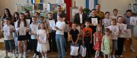 Zdjęcie grupowe w sali oficjalnej. Duża grupa dzieci i młodzieży w różnym wieku stoi w dwóch rzędach i pozuje do zdjęcia, trzymając w rękach pamiątkowe dyplomy oraz białe upominki w torbach. W centralnej części, z tyłu, stoją trzej dorośli mężczyźni – dwaj w garniturach oraz jeden w piaskowym mundurze strażackim. W tle widoczne są tablice z pracami plastycznymi oraz duże, pionowe wstęgi w barwach flag: żółto-niebieskiej, biało-czerwonej i błękitnej z herbem.