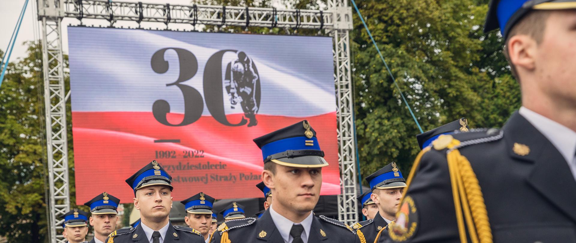 Maszeruje kompania reprezentacyjna, za nimi na telebimie wyświetlona flaga RP z napisem okolicznościowym 30 lecia PSP