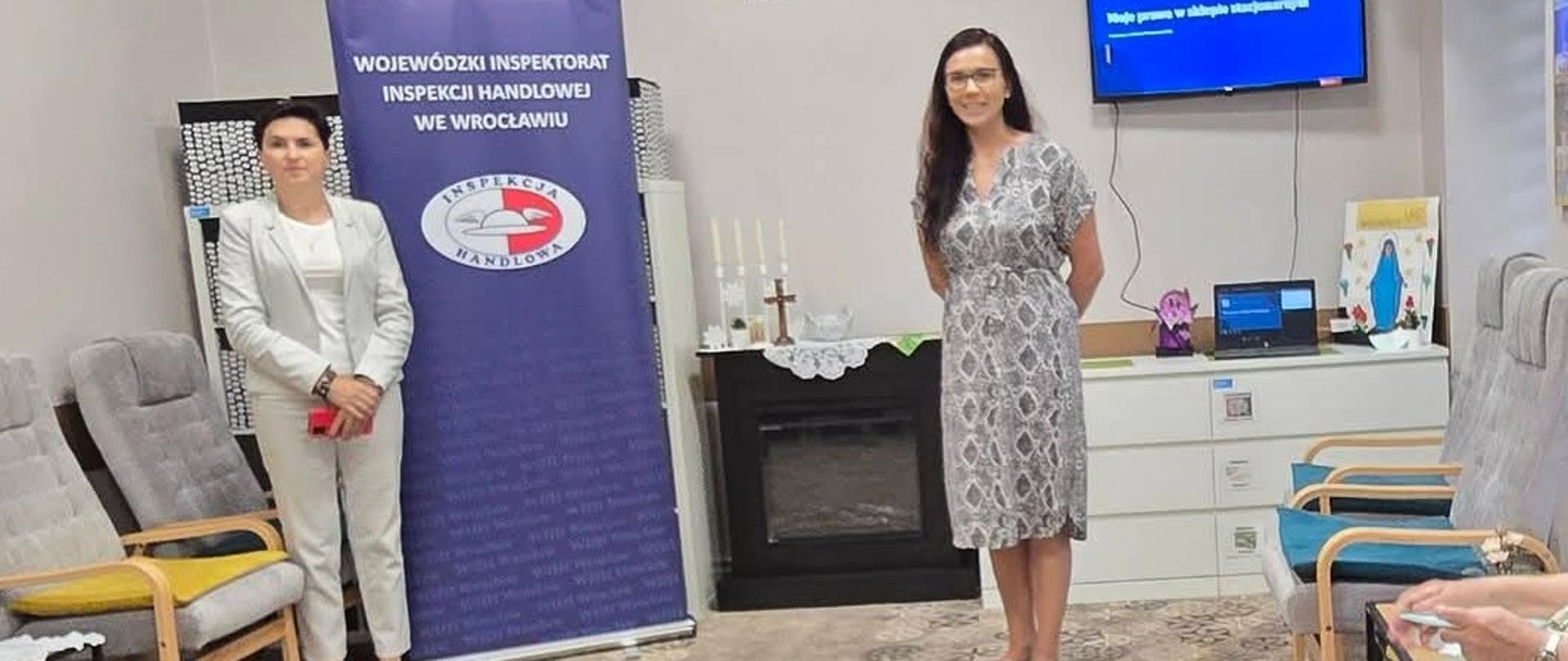 Dwie kobiety - prelegentki stojące na środku dużego pokoju. Po obu stronach pokoju stoją fotele w szarych odcieniach. Za kobietami znajduje się po lewej stronie duży banner Inspekcji Handlowej, centralnie czarny kominek i po prawej stronie telewizor. W centralnym miejscu na ścianie w tle ikona Matki Bożej. 