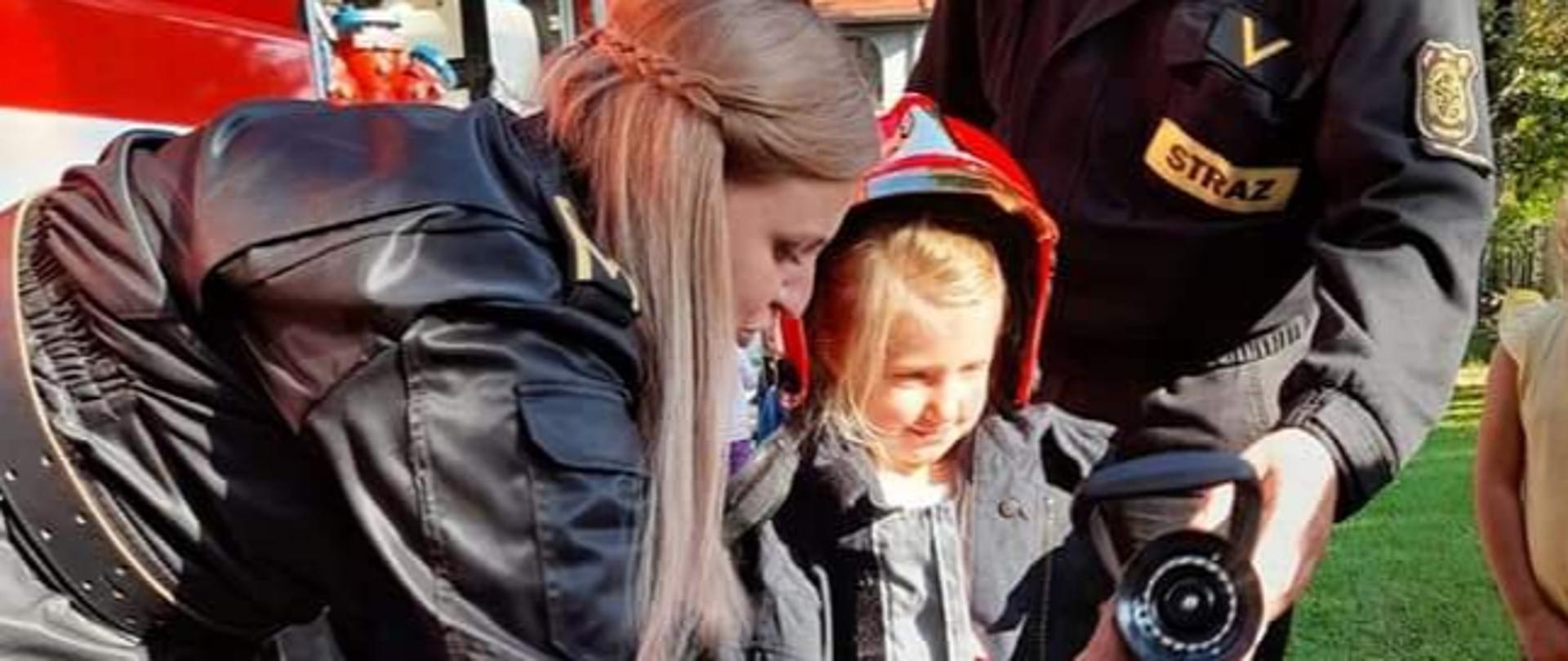 strażacy pomagają małej dziewczynce ubranej w kurtkę strażacka, w tle czerwony wóz strażacki