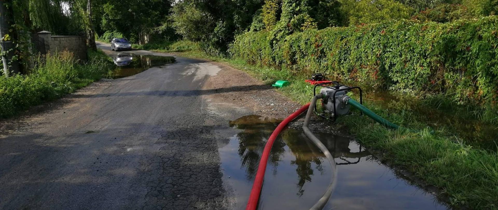 Na ulicy widać zalaną drogę. Pompy strażackie wypompowują wodę. Jest pogodny dzień.