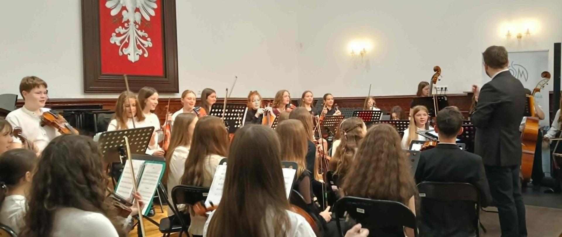 Orkiestra smyczkowa złożona z uczniów gra pod kierunkiem dyrygenta w Sali Królewskiej, w tle herb Polski i freski z portretami królów polskich.