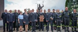 Zdjęcie zrobione na tle figurki św. Floriana. Na pierwszym planie pozuje grupa strażaków w umundurowaniu wyjściowym i koszarowym.