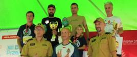 Czterech mężczyzn i dziecko w strojach sportowych stoi na podium, przed nimi stoi trzech mężczyzn, dwóch stojących po bokach ubranych jest w mundury straży pożarnej, stojący w środku jest w stroju sportowym