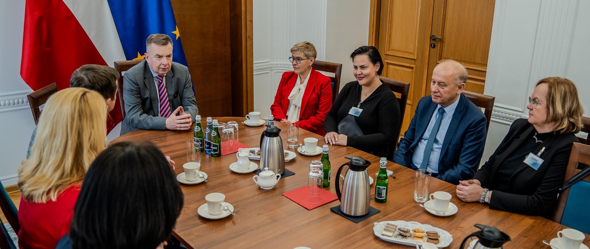 Zdjęcie z góry pod kątem, w sali przy podłużnym stole siedzą ludzie, u szczytu stołu minister Wieczorek, za nim pod ścianą flagi Polski i UE.