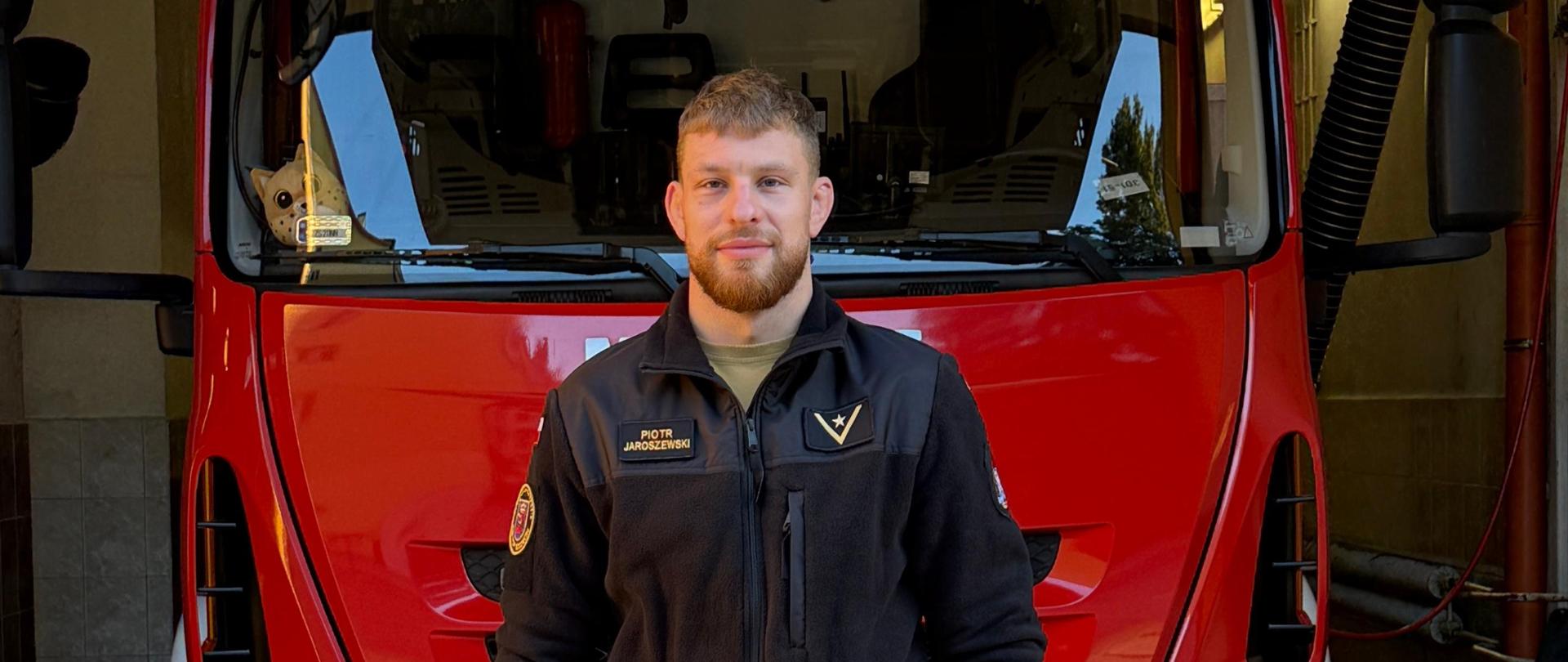Zdjęcie przedstawia strażaka stojącego na tle czerwonego wozu strażackiego. Ma założoną czarną bluzę z naszywką Komendy Miejskiej Państwowej Straży Pożarnej w Szczecinie oraz polską flagą na ramieniu. Strażak ma skrzyżowane ręce, na nadgarstku widoczny zegarek. Twarz osoby została zamazana ze względów prywatności. W tle, na szybie pojazdu, znajduje się maskotka kota.