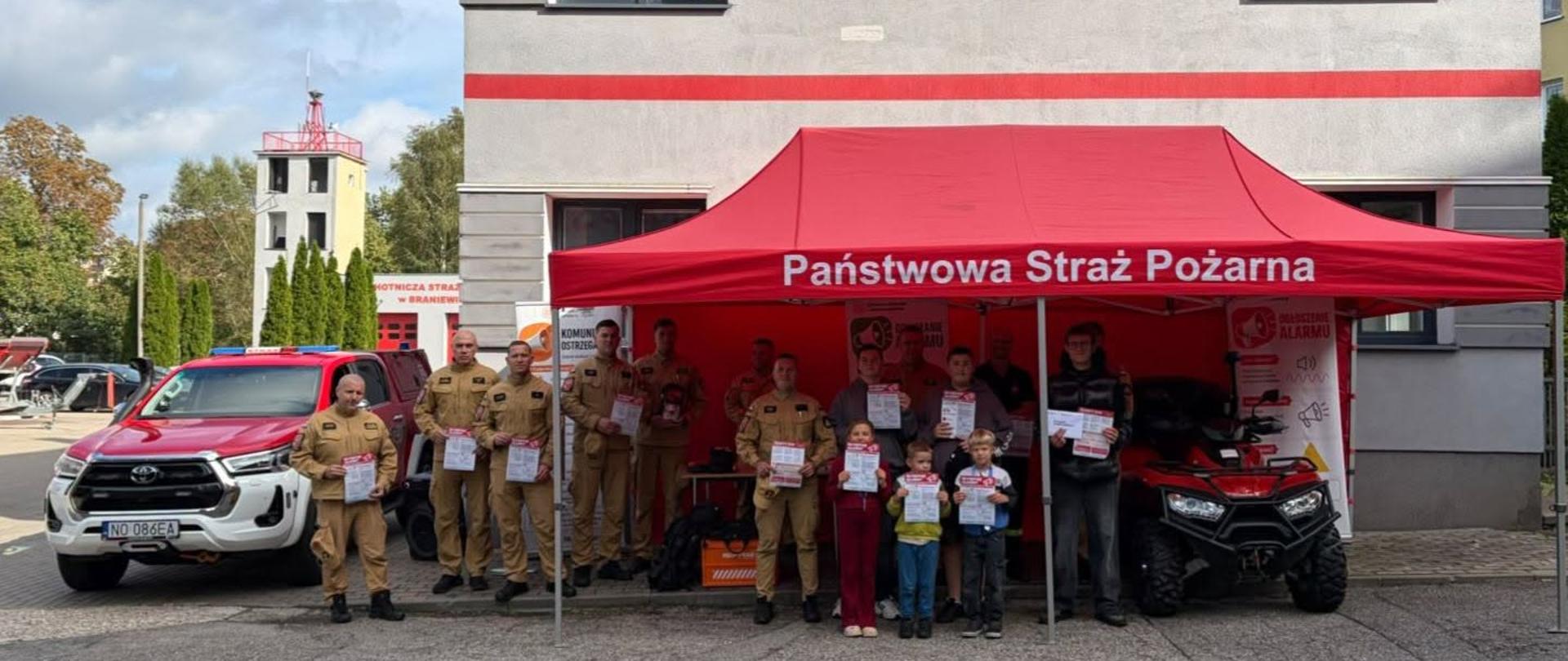 Na fotografii przed budynkiem Komendy Powiatowej Państwowej Straży Pożarnej w Braniewie stoi grupa strażaków oraz kilkoro dzieci. Większość strażaków ubrana jest w piaskowe mundury służbowe i trzyma w dłoniach materiały związane z treningiem reagowania. Nad uczestnikami rozstawiony jest duży czerwony namiot z białym napisem „Państwowa Straż Pożarna”.
