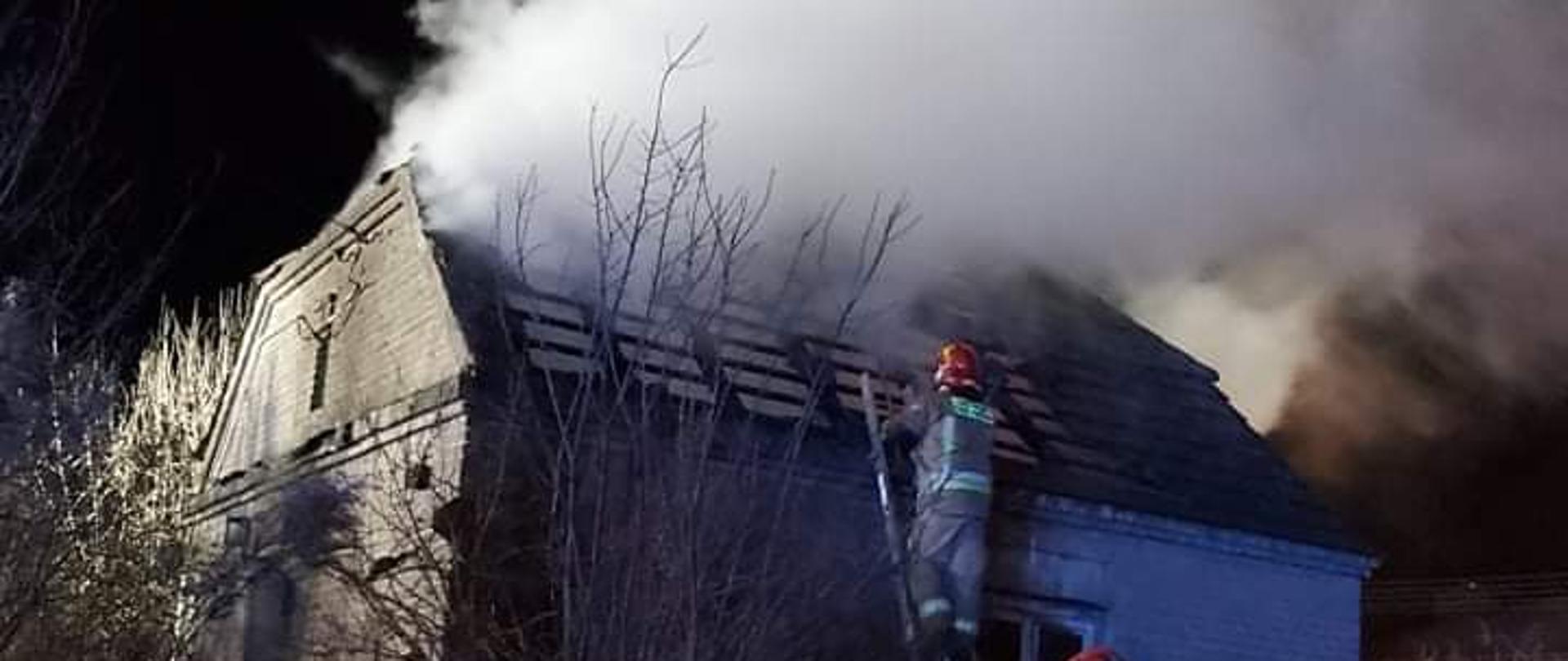 Na zdjęciu w porze nocnej widać dwóch strażaków, jeden na drabinie, drugi zaś przy niej, którzy gaszą pożar domu mieszkalnego, jednorodzinnego w Ostrowcu Św. Na zdjęciu widać wydobywający się dym z dachu budynku.