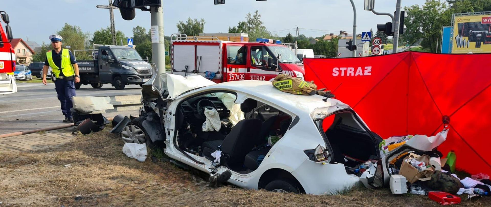 Uszkodzony samochód w rowie na tle pojazdu strażackiego i parawanu.