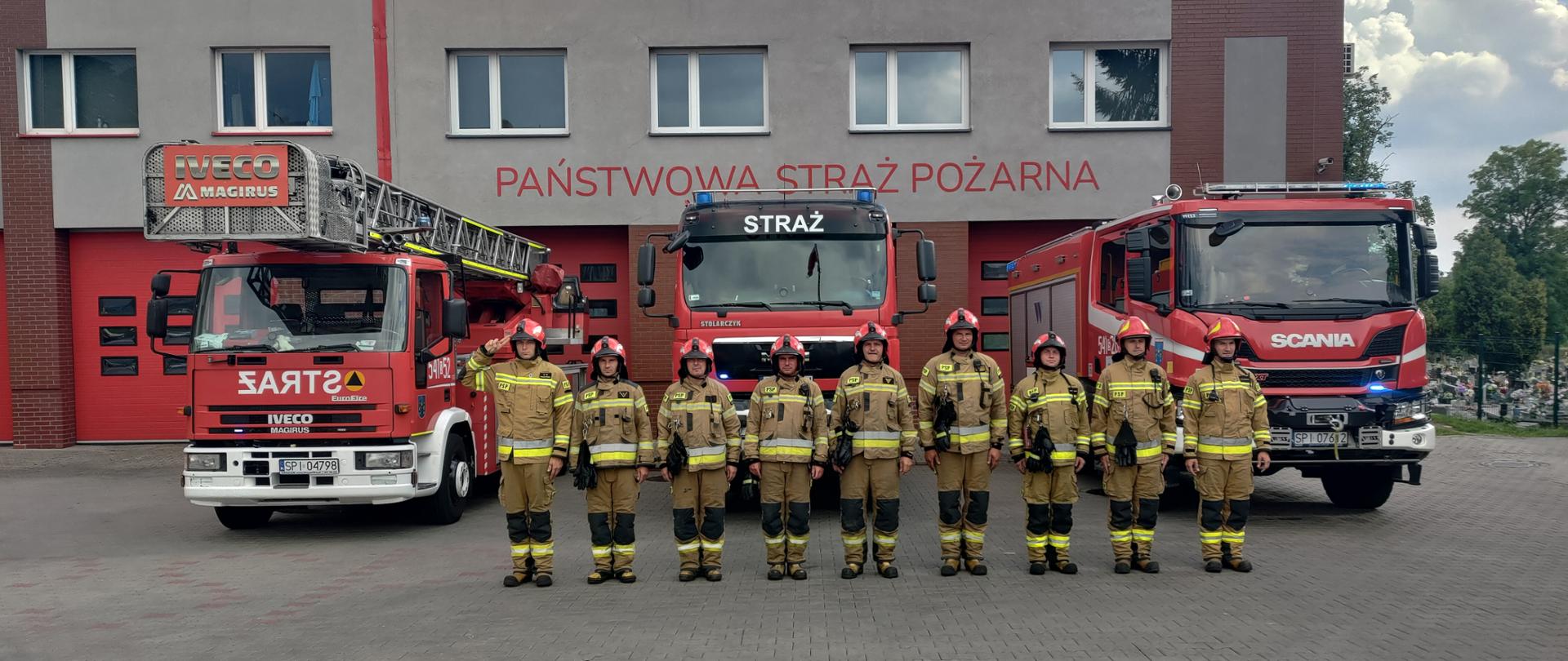 Zdjęcie przedstawia 9 strażaków ubranych w ubrania specjalne stojących w szeregu na tle samochodów pożarniczych