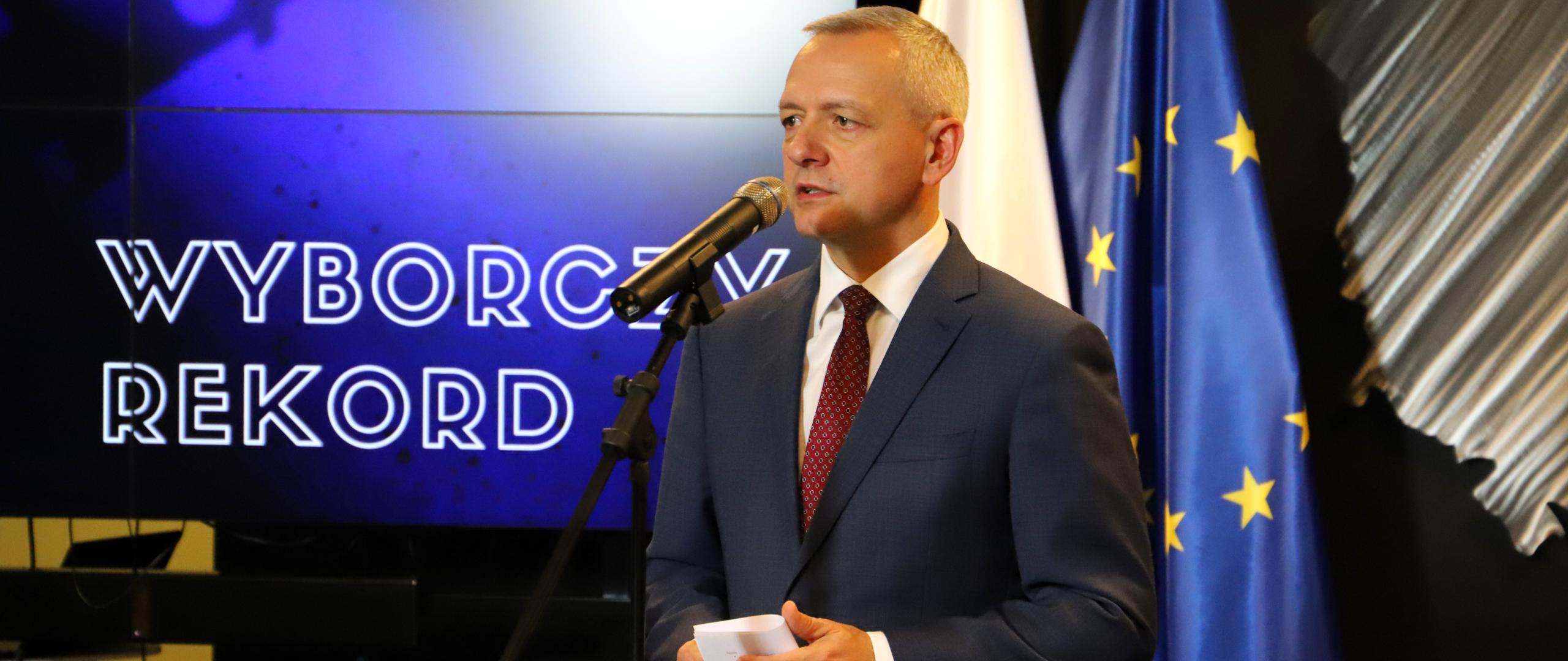 Minister Marek Zagórski stoi przy mikrofonie, w tle flagi polska i unijna oraz ekran z napisem wyborczy rekord