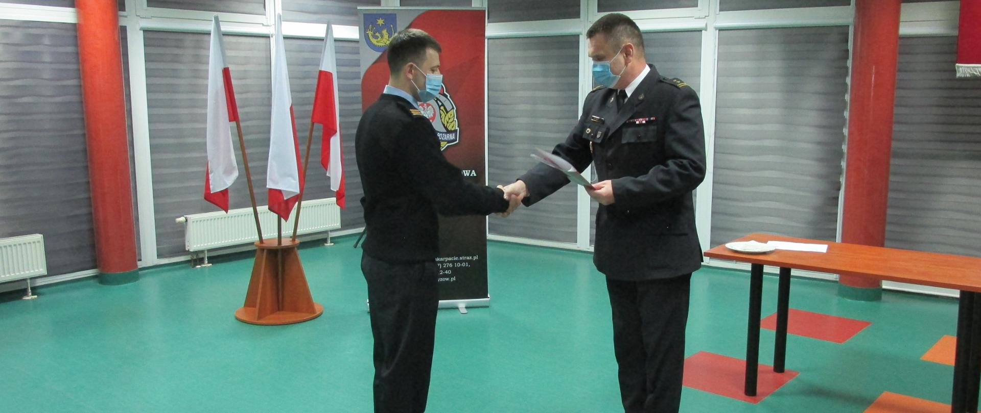 Na zdjęciu Komendant Powiatowy Państwowej Straży Pożarnej w Strzyżowie brygadier Rafał Rytuna w umundurowaniu wyjściowym oraz starszy sekcyjny Kacper Pirga w umundurowaniu służbowym podaja sobie prawe ręce. Komendant w lewej ręce trzyma teczkę. Obaj mają zasłanięte twarze maseczką. W tle trzy Flagi Polski oraz baner Komendy Powiatowej Państwowej Straży Pożarnej w Strzyżowie. Po prawej stronie widać stół.