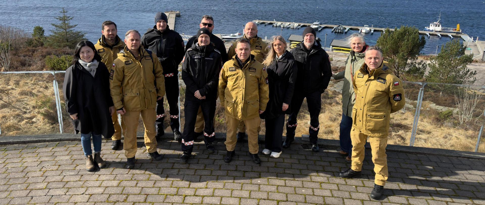 Grupa strażaków i przedstawicieli instytucji stoi na zewnątrz nad brzegiem wody, pozując do wspólnego zdjęcia na tle przystani i skalistego krajobrazu.
