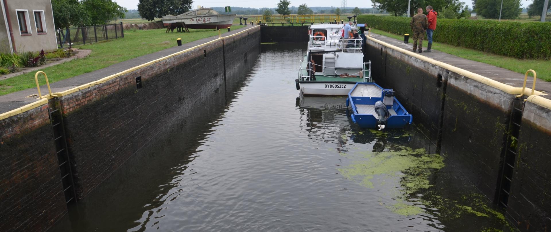 Nakło Zachód - Śluza nr 8 drogi wodnej Wisła-Odra, która jest elementem międzynarodowej drogi wodnej E-70. Na pierwszym planie komora śluzy napełniana wodą. Wrota śluzy zamknięte. Trwa śluzowanie. W komorze statek pasażerski o nazwie Bydgoszcz. Na pokładzie statku wodniacy podróżujący rzeką Noteć do kolejnej śluzy. Po prawej stronie śluzy dwóch mężczyzn operatorów śluzy. 