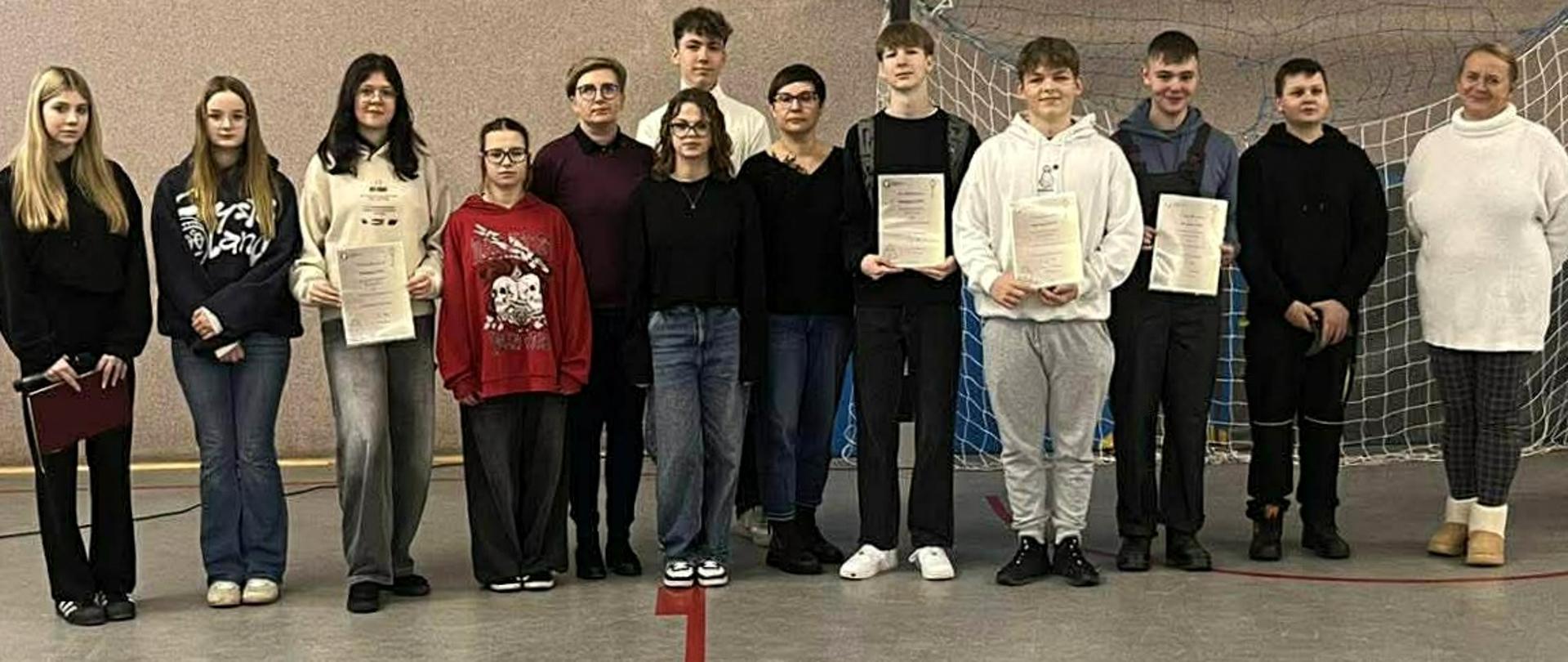Grupa uczniów z nauczycielami w sali gimnastycznej pozuje do zdjęcia, niektórzy trzymają w rękach dyplomy.