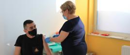 Strażak podczas podania szczepionki przeciw COVID-19. Funkcjonariusz siedzi w gabinecie zabiegowym a pielęgniarka podaje zastrzyk w lewe ramie strażaka. Zarówno strażak jak i pielęgniarka mają zasłonięte usta i nos maseczkami ochronnymi. 