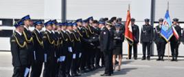 Na zdjęciu strażacy ustawieni w szeregu, przyjmują awanse na wyższe stopnie służbowe wręczane przez Śląskiego Komendanta Wojewódzkiego Państwowej Straży Pożarnej i Komendanta Miejskiego Państwowej Straży Pożarnej w Bytomiu. W tle widoczne poczty sztandarowe i budynek komendy.