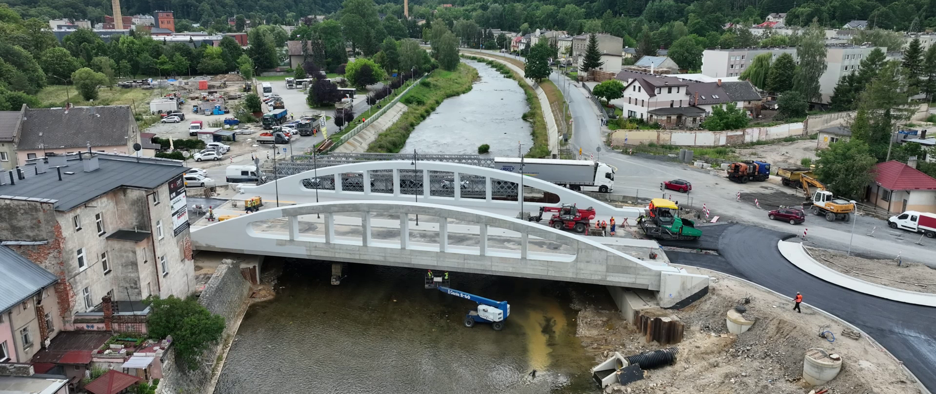 Widok z lotu ptaka na nowy most nad rzeką w trakcie realizacji inwestycji drogowej. Na moście i dojazdach pracują maszyny budowlane oraz ekipy drogowe. Wokół widoczne są zabudowania miejscowości, biegnąca wzdłuż rzeki droga oraz zalesione wzgórza w tle pod zachmurzonym niebem.