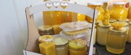 Na stole i w skrzynce stoją słoiki miodu, świece woskowe i zapakowane plastry miodu