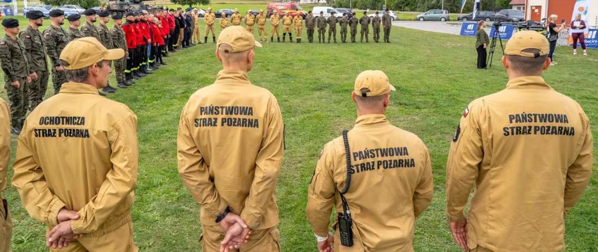 Szeroki kadr przedstawiający uczestników manewrów ustawionych podczas oficjalnej zbiórki. Widać przedstawicieli różnych służb i formacji, w tym PSP, OSP oraz żołnierzy. Na pierwszym planie znajdują się strażacy PSP i OSP, a cały kadr dobrze pokazuje skalę przedsięwzięcia i współpracę między służbami.