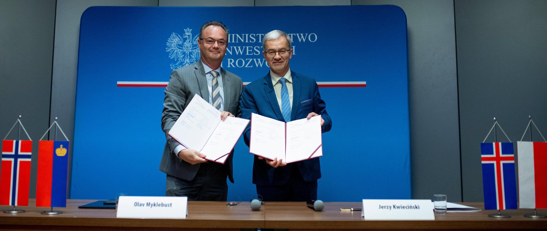 Na zdjęciu ambasador Olav Myklebust i minister Jerzy Kwieciński pokazujący otwarte teczki z podpisanymi umowami. Z przodu stół, na nim proporczyki: Norwegii, Liechtensteinu, Islandii i Polski oraz koziołki z imionami i nazwiskami ambasadora i ministra. Z tyłu granatowa ścianka z orłem i napisem "ministerstwo inwestycji i rozwoju"