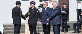 Moment wręczania odznaczeń lub gratulacji. Strażak w mundurze galowym podaje rękę oficerowi straży. Obok stoją dwaj mężczyźni w ciemnych garniturach, obserwując ceremonię. Wydarzenie odbywa się na zewnątrz przy jasnym budynku.